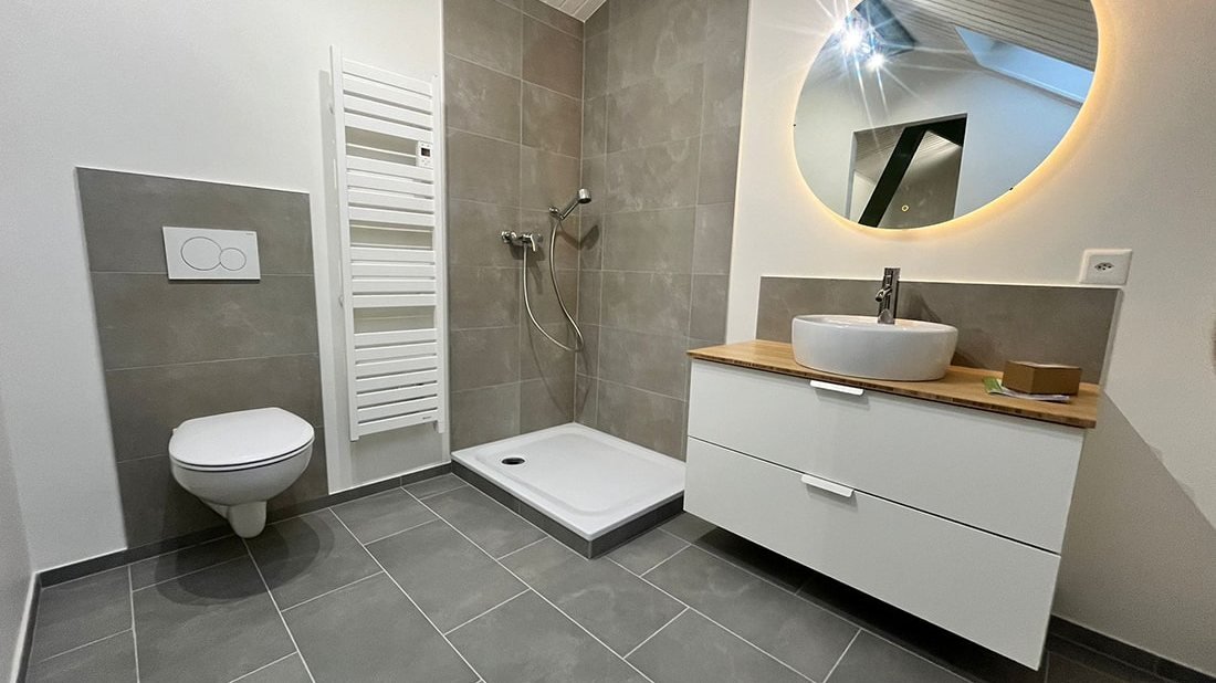 Salle de bain neuve : douche carrelée gris, sèche-serviettes, WC suspendu, miroir rond rétroéclairé et meuble-vasque